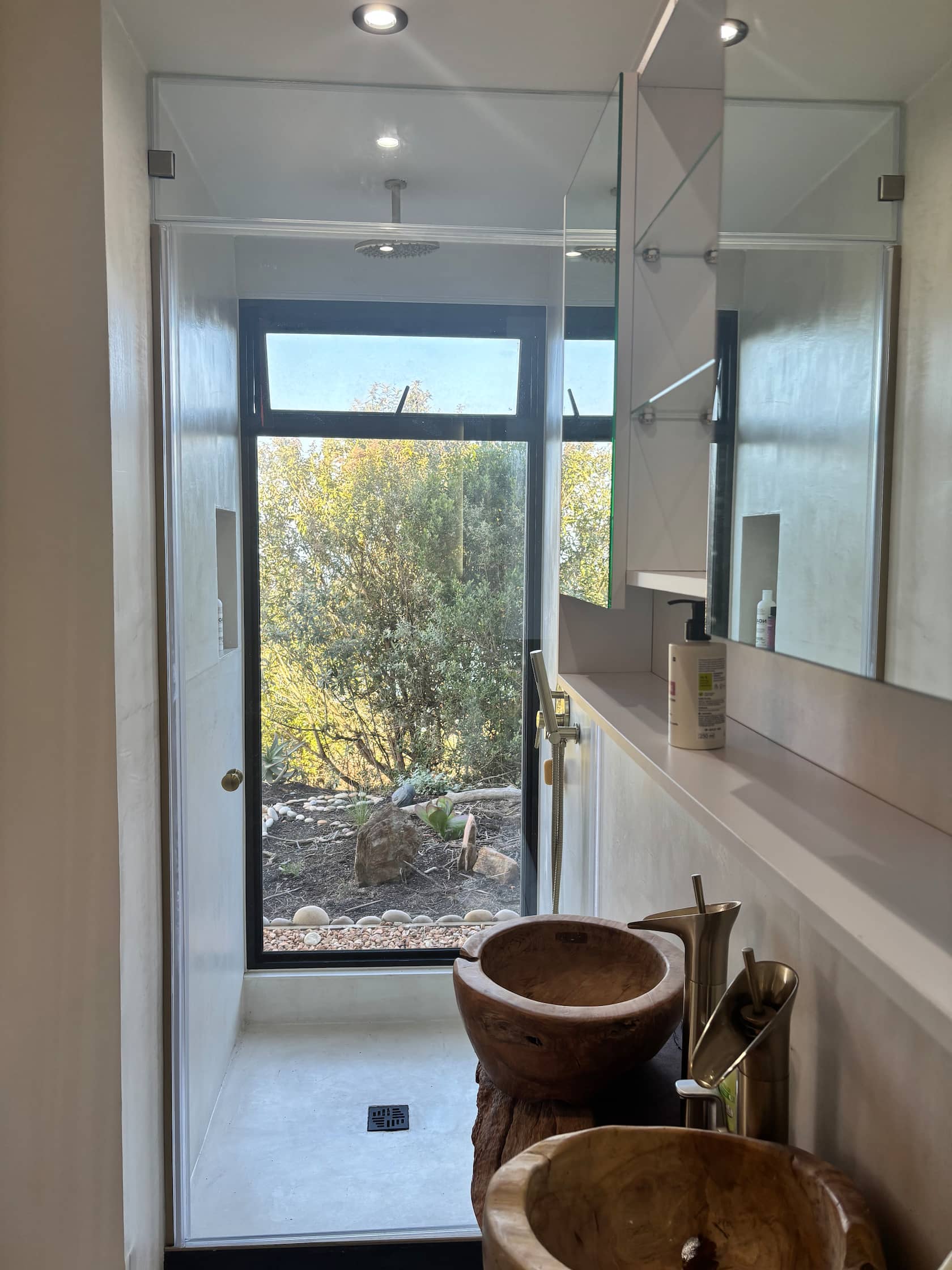 Walk-in rainfall shower and wooden vessel basins beside a garden window